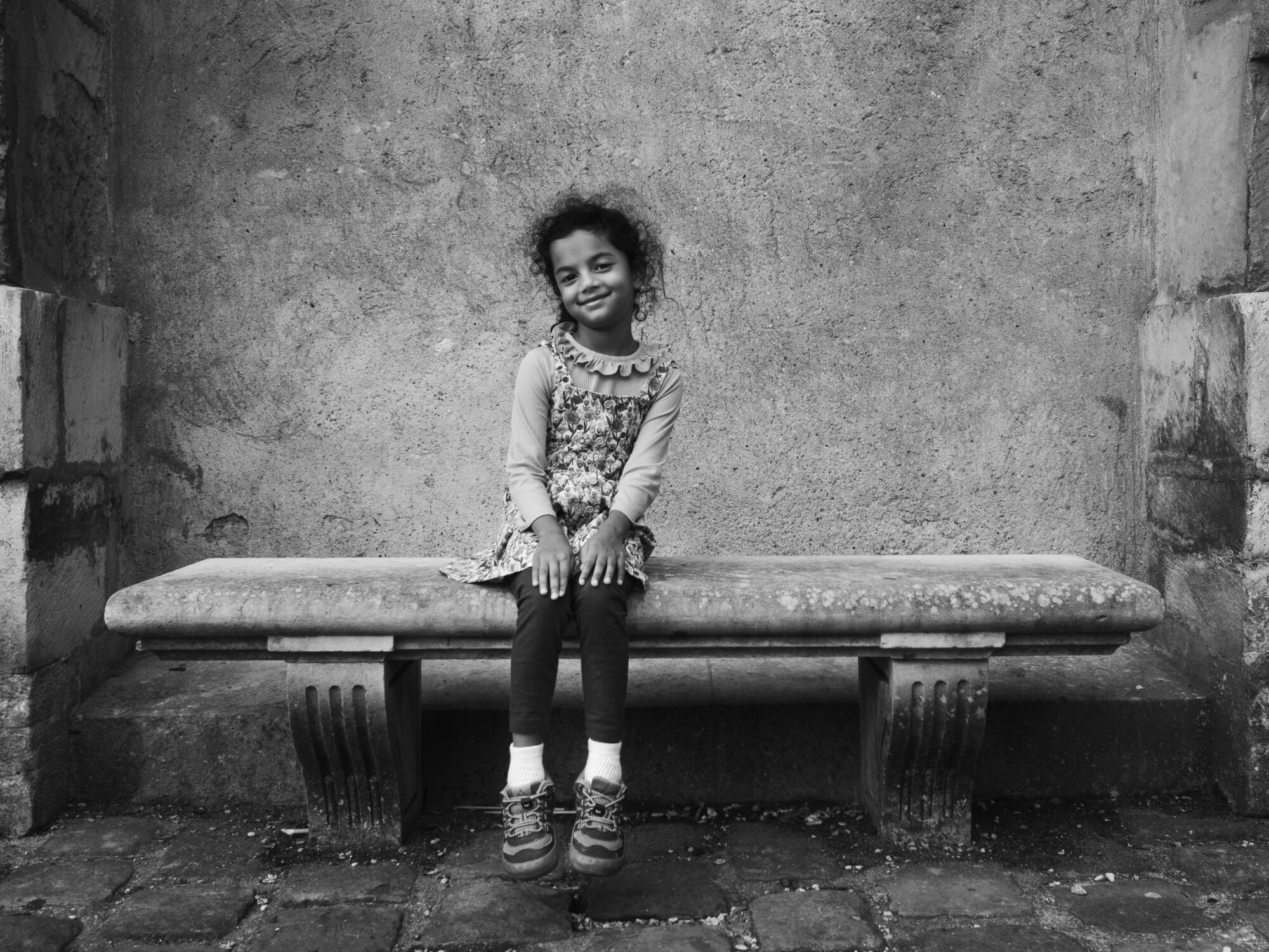Little girl sitting on a stone bench. She has her hands on her knees and is smiling.