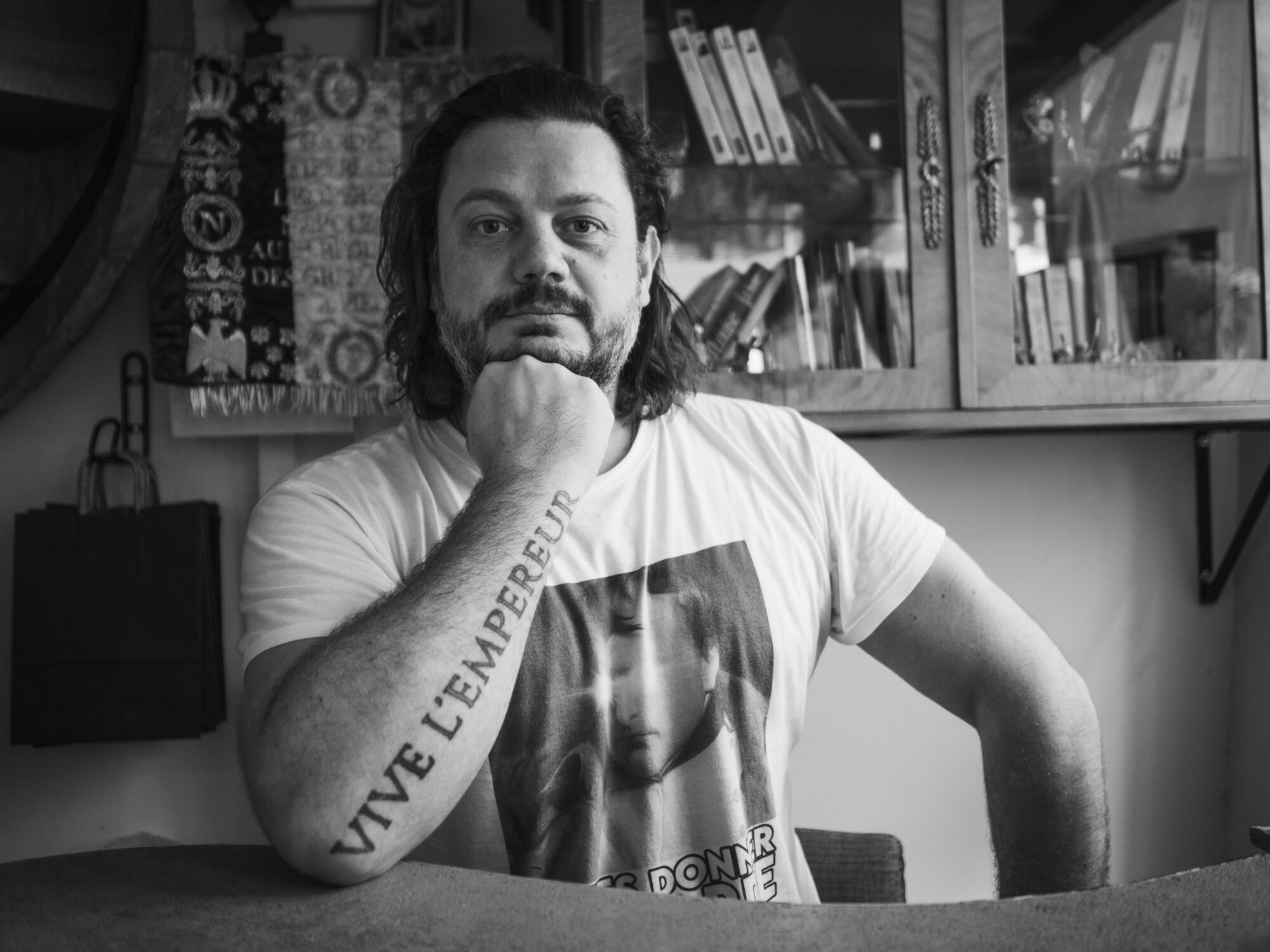 A middle aged man leaning on his arm showing a tattoo that says Vive L'Empereur